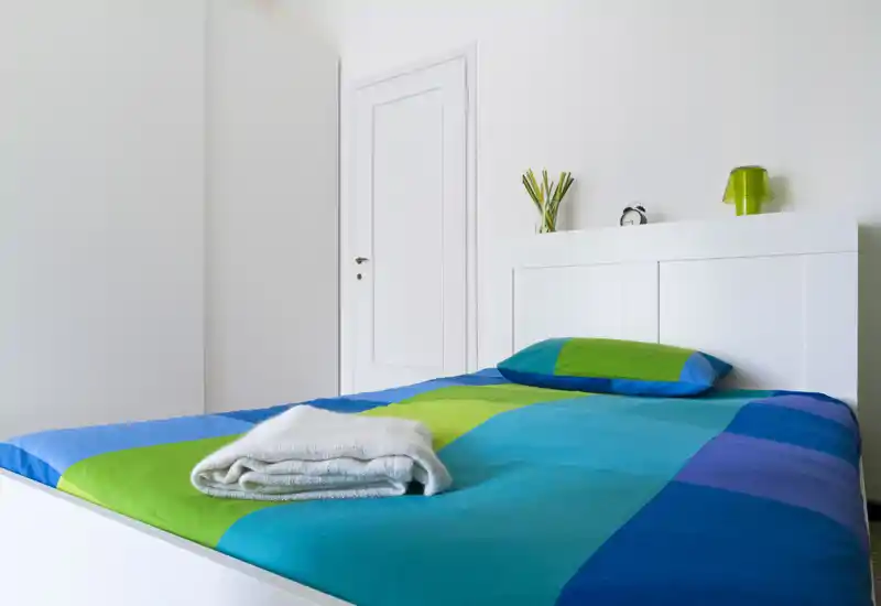 Camera da letto moderna e luminosa con testiera bianca e biancheria dai toni blu-verdi. Il letto è ben rifatto, con asciugamani piegati e una mensola con lampada e orologio.