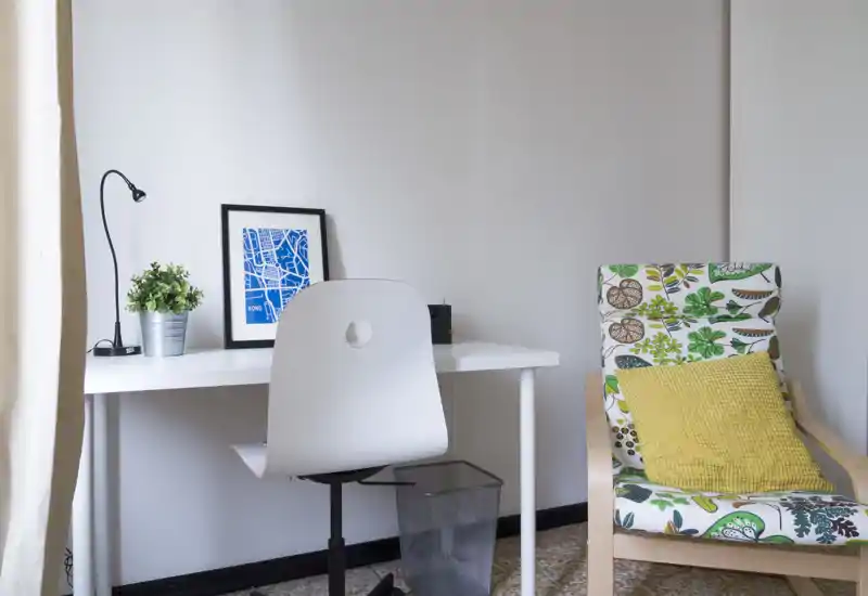 Angolo soggiorno luminoso e ordinato con una scrivania bianca e una sedia moderna accanto a una poltrona con fantasie e cuscino giallo; arredamento minimal con lampada da scrivania, stampa incorniciata e pianta.