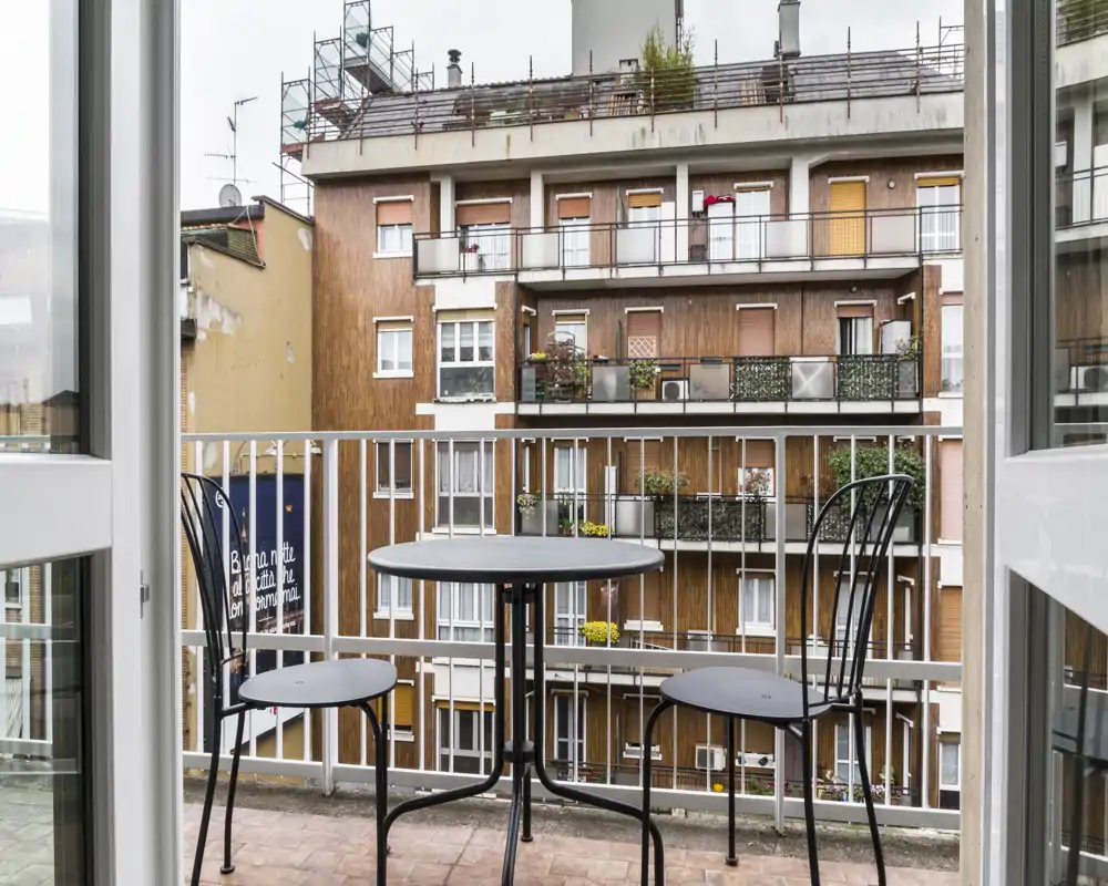 Piccolo balcone urbano con tavolo rotondo in metallo e due sedie, affacciato sulle facciate dei palazzi vicini — ideale per un caffè mattutino in città.