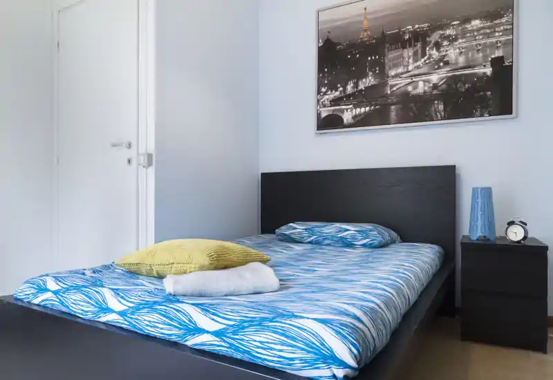 Camera da letto luminosa e ordinata con letto matrimoniale coperto da biancheria blu con motivi, cuscini decorativi e comodino con lampada e sveglia.