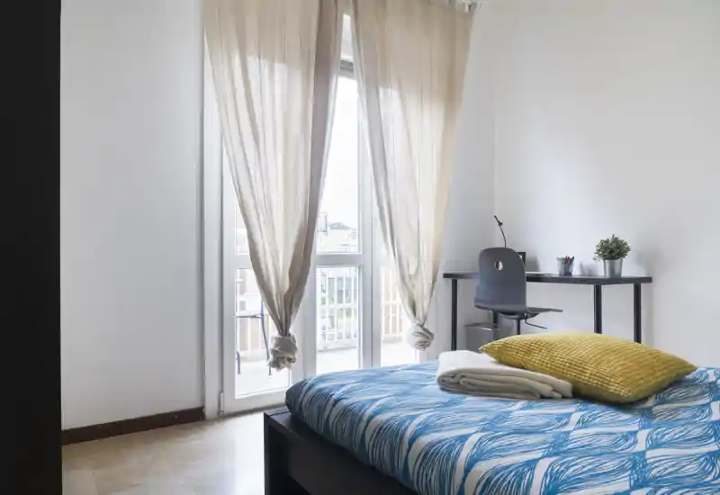 Camera da letto luminosa e moderna con letto matrimoniale con copriletto blu, cuscino color senape e lenzuola piegate. Ampia finestra con tende leggere che illumina l'ambiente; una scrivania semplice e sedia formano un angolo studio.