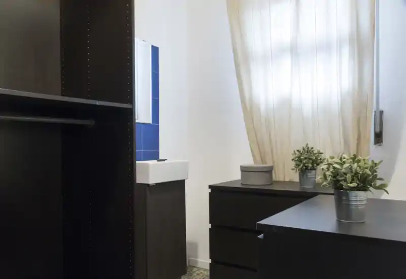Angolo di ripostiglio/guardaroba in camera con armadi scuri e cassettiere basse, luce naturale soffusa dalla finestra con tendina e piccole piante in vaso come decorazione.