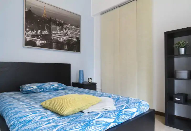 Camera da letto moderna e luminosa con un grande letto con copertura blu a fantasia, cuscino decorativo giallo, comodino e scaffale — spazio accogliente e curato, ideale per annunci.