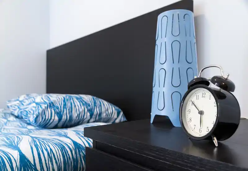 Primo piano di una camera da letto moderna con comodino, sveglia, lampada decorativa blu e letto ordinato con biancheria a fantasia blu. Composizione pulita e stile contemporaneo con luce naturale morbida.