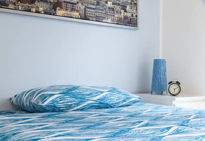 Bright, modern bedroom close-up with a blue patterned duvet and matching pillow on a neatly made bed; a bedside table holds a small lamp and alarm clock.