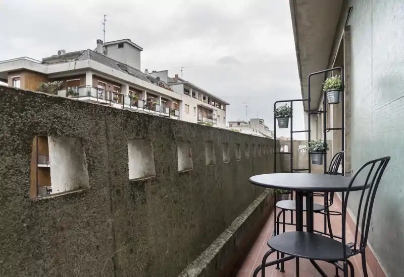 Sunny-feel urban balcony with a small round table, two metal chairs and vertical plant shelves — ideal for a morning coffee while overlooking neighboring apartments.