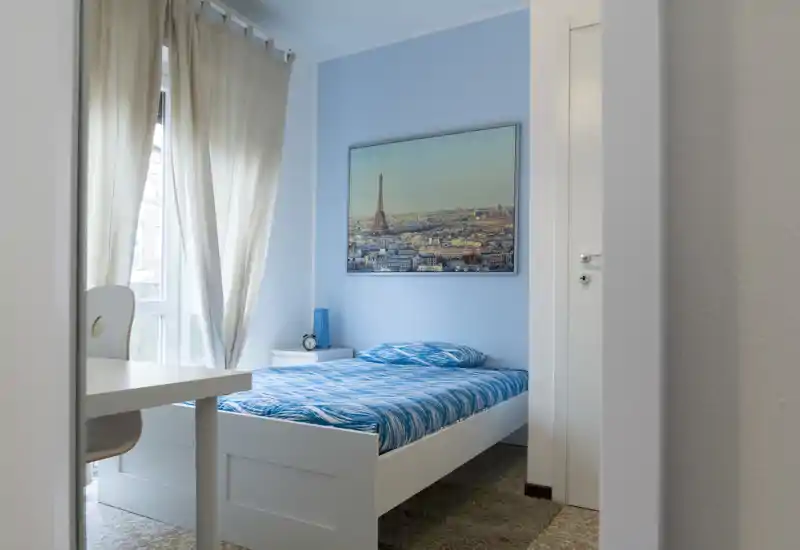 Habitación individual luminosa y ordenada con cama de estructura blanca, ropa de cama azul con estampado, mesita de noche y ventana grande con cortinas claras; luz natural suave y cuadro de ciudad en la pared.