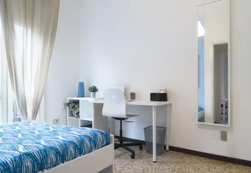 Dormitorio luminoso y minimalista con cama de estampado azul, escritorio blanco y silla junto a la ventana. Luz natural, decoración sencilla y espejo de cuerpo entero crean un espacio fresco y tranquilo, ideal para alquileres de corta estancia.