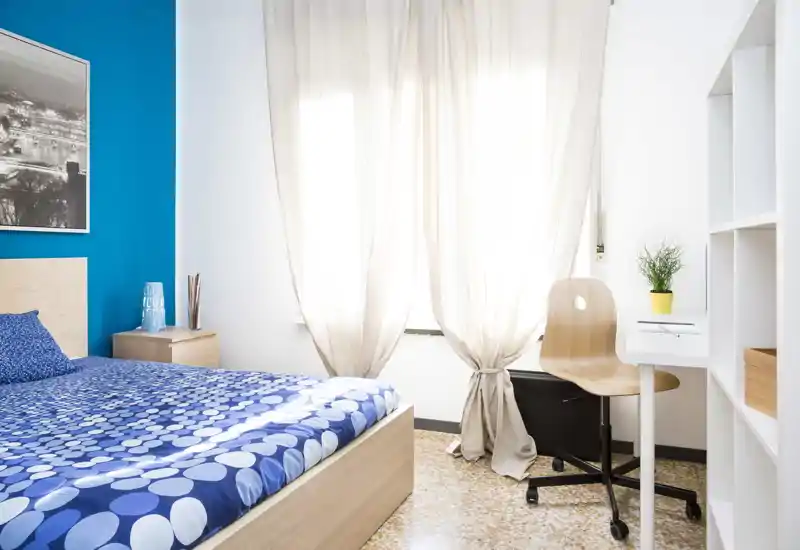Chambre lumineuse et bien rangée avec un mur bleu et une couette bleue à motifs. On y trouve une table de chevet, une grande fenêtre avec voilages et un petit espace de travail avec chaise.