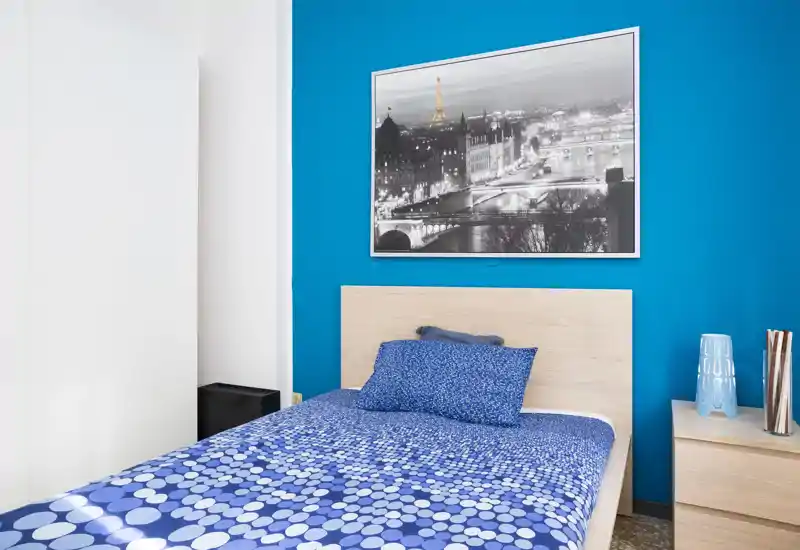 Chambre moderne et lumineuse avec un lit simple habillé d’un linge de lit bleu à motifs, une tête de lit en bois clair et une table de chevet assortie; mur bleu vif avec une photo urbaine encadrée.