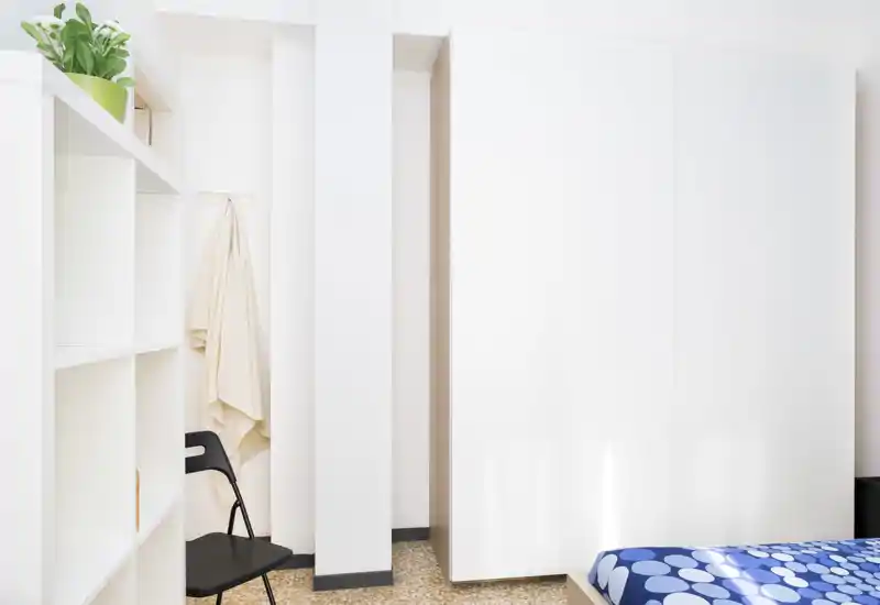 Minimal bright bedroom corner with a patterned blue bedspread, white wardrobe and shelving; clean, airy styling ideal for rental photos.