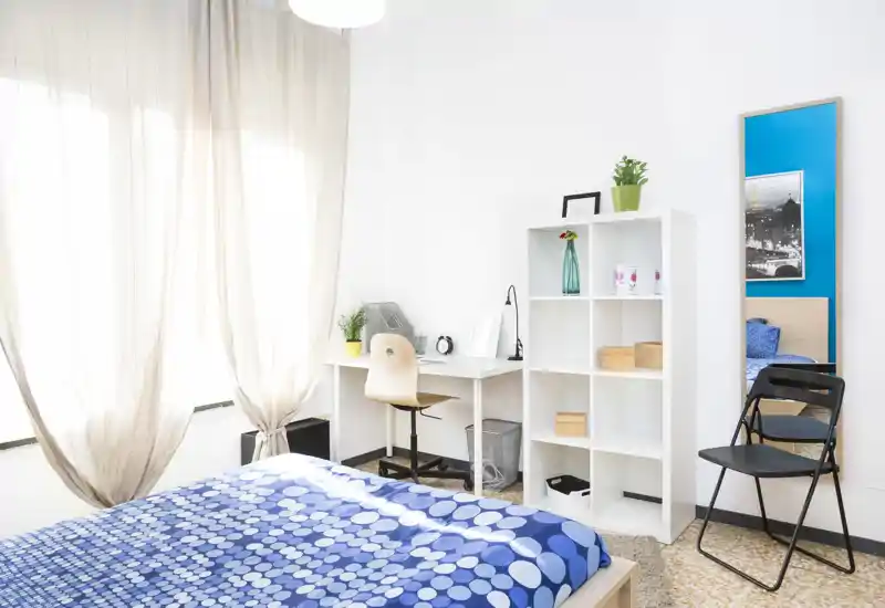 Chambre moderne et lumineuse avec couette bleue à motifs, espace bureau près de la fenêtre et étagères ouvertes — aérée, bien éclairée et soignée.