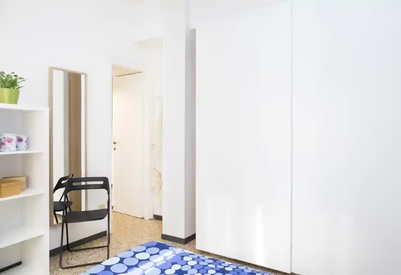 Bright, minimal bedroom corner with a patterned blue bedcover, wardrobe with sliding white doors and a full-length mirror beside a simple shelf and folding chair.