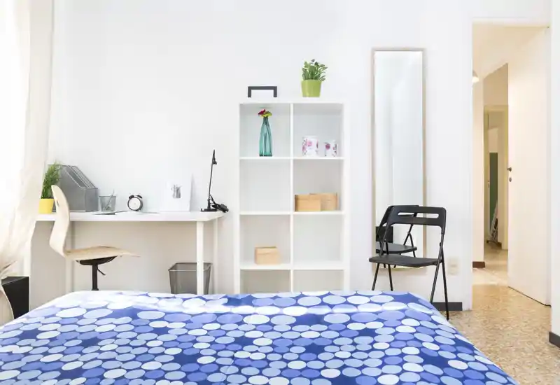 Chambre lumineuse et minimaliste avec une couette bleue à motifs au premier plan, un bureau rangé et une étagère cubique contre un mur blanc, et une grande glace près de la porte.