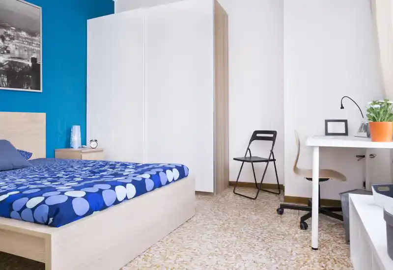 Camera da letto moderna e luminosa con letto matrimoniale con copriletto blu a fantasia, comodino e armadio; postazione di lavoro con scrivania e sedia a destra.