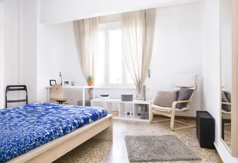 Camera da letto luminosa e minimale con struttura letto in legno, copriletto blu a motivi, scrivania vicino alla finestra e una poltrona accogliente accanto allo specchio a figura intera.