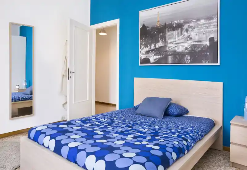 Camera da letto moderna e luminosa con letto matrimoniale con biancheria blu a fantasia, testiera in legno, comodino e una grande foto su una parete accentuata in blu vivo.