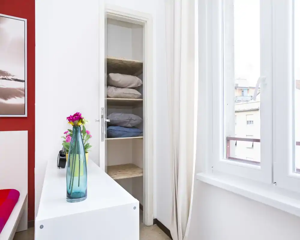 Bright, modern bedroom corner with a partial view of the bed, a simple white dresser topped with a blue glass vase and flowers, shelving with folded linens and a large window letting in natural light.