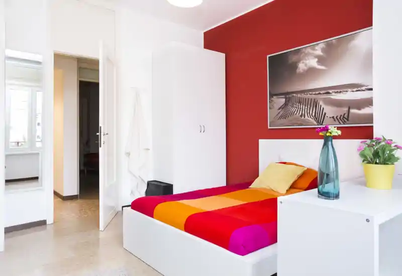 Bright, modern single bedroom with a colorful bedspread, simple white furniture and decorative plants; strong natural light and bold red accent wall create a lively, inviting atmosphere.