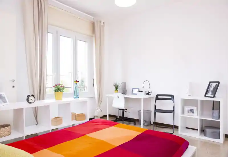 Bright, modern bedroom with a colorful bedspread, large window letting in abundant natural light, desk workspace and minimalist shelving—comfortable and well-styled for rental listings.