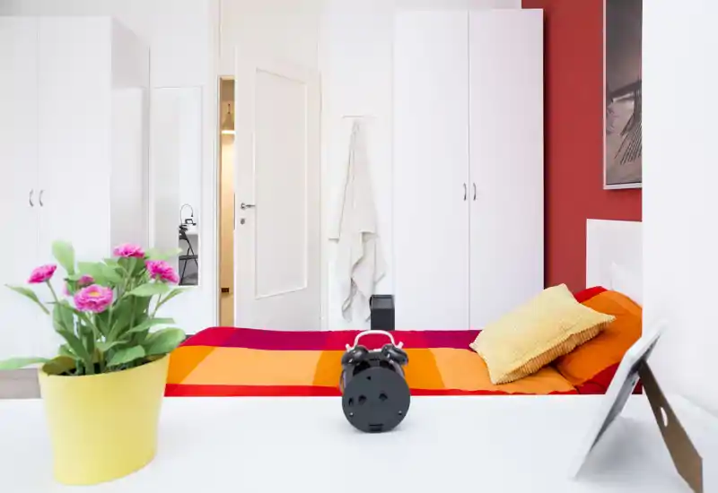 Bright, colorful bedroom with a neatly made bed in warm orange and red tones, a textured cushion and white wardrobes; a cheerful, well-lit space ideal for rental photos.