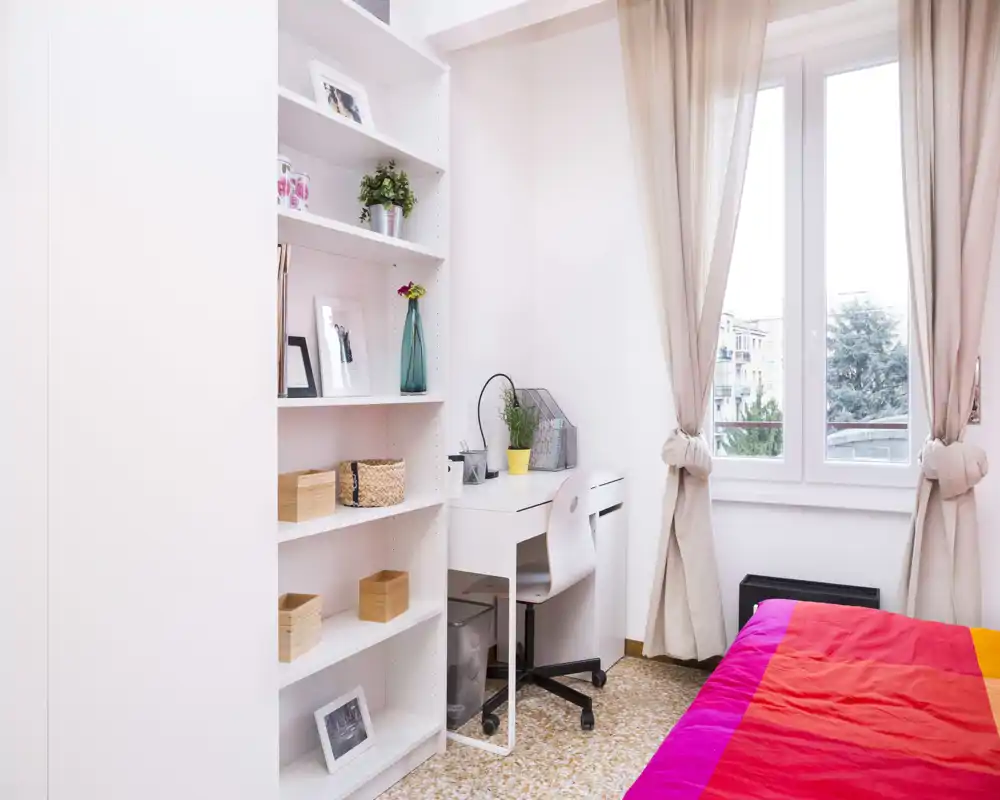 Bright, cozy bedroom corner with a colorful bedspread, a small desk by the window and organized shelving — ideal for a single occupant or student.
