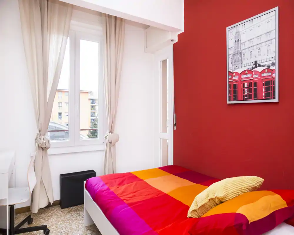Bright, colorful bedroom with a large bed, vivid red accent wall and natural light from a window; simple desk and chair included.