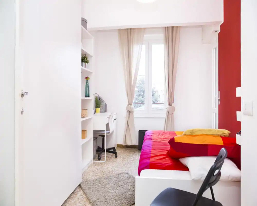 Bright, compact bedroom with a colourful double bed, natural light from a window and a small work desk beside built-in shelving.
