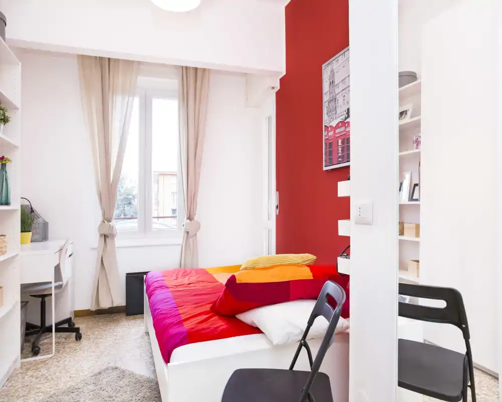 Bright, colorful bedroom with a double bed dressed in vibrant red and orange linens, a window with neutral curtains providing natural light, shelving and a small desk — ideal for a cheerful rental listing.