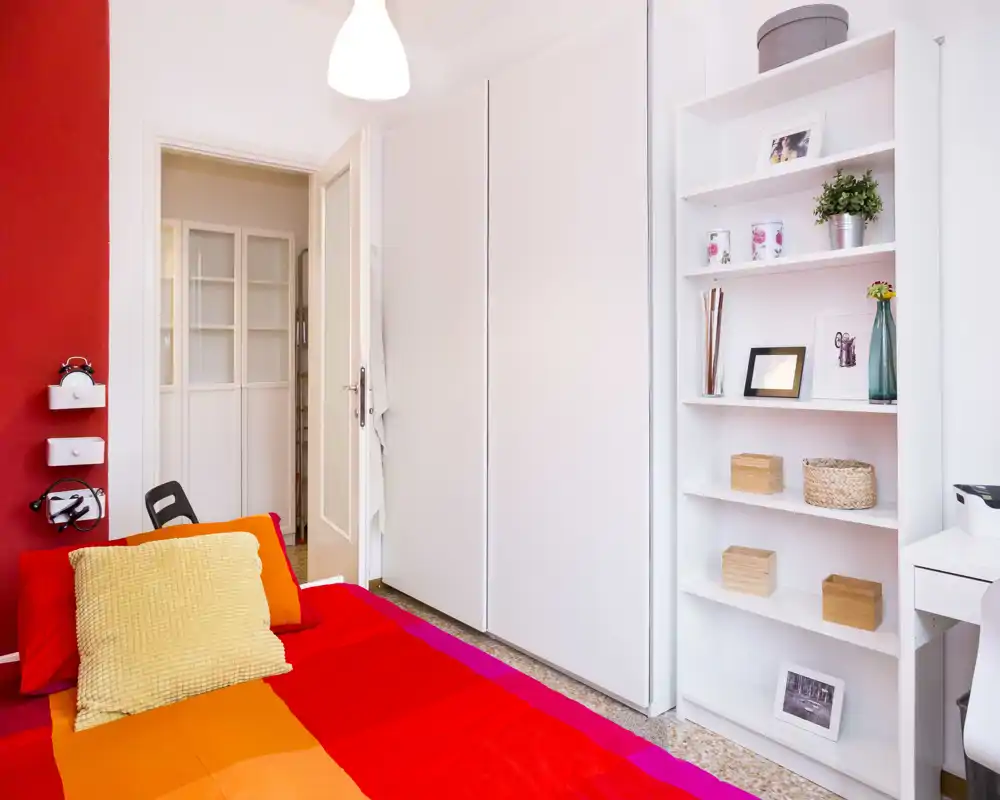 Bright, colorful bedroom with a large red-and-orange bed in the foreground, a tall white wardrobe and open shelving with decorative items. The room is well lit and styled for a cozy, modern feel.