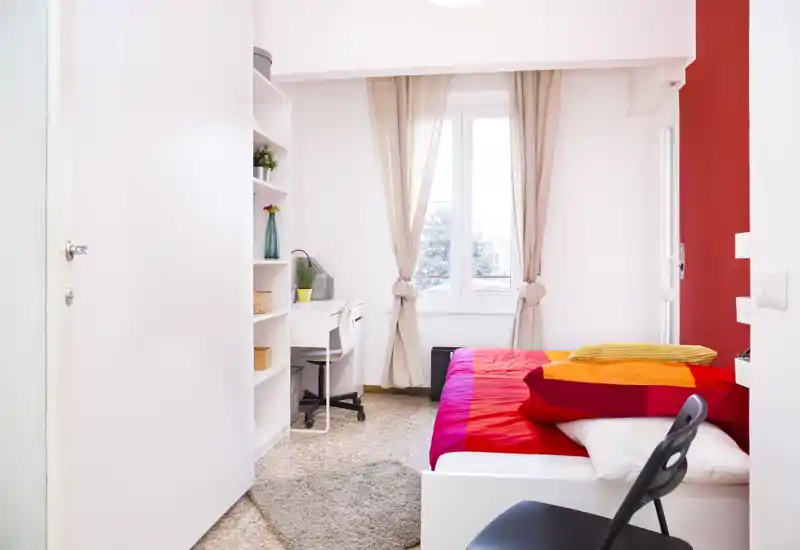 Bright, tidy bedroom with a white bed frame and bold red-orange bedding, a desk and shelving beside a window that fills the room with natural light.