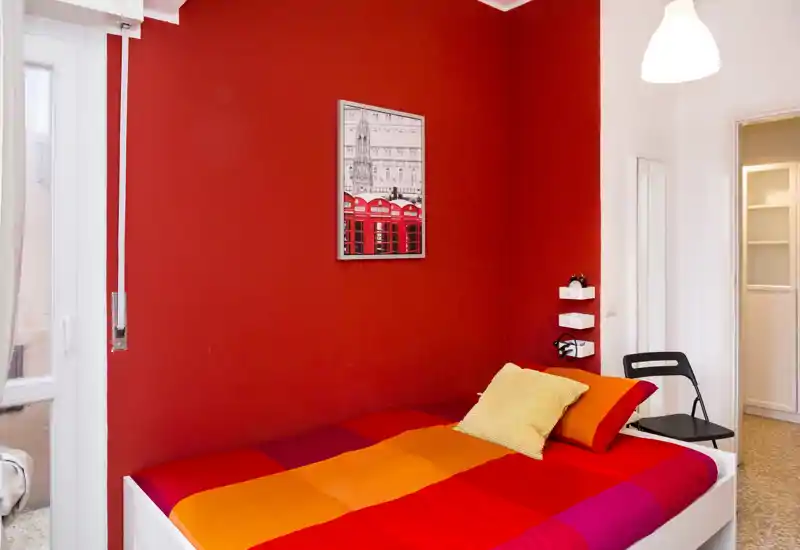 Bright, colorful bedroom with a single bed against a vivid red wall, warm orange-red bedding and a decorative pillow. The room feels cozy and well lit, ideal for a single traveler or student.