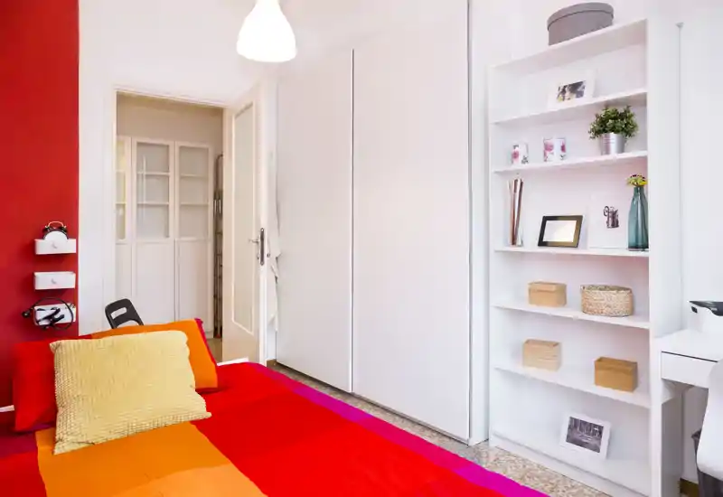 Bright, colorful bedroom with a large red-and-orange bed in the foreground, a tall white wardrobe and open shelving with decorative items. The room is well lit and styled for a cozy, modern feel.