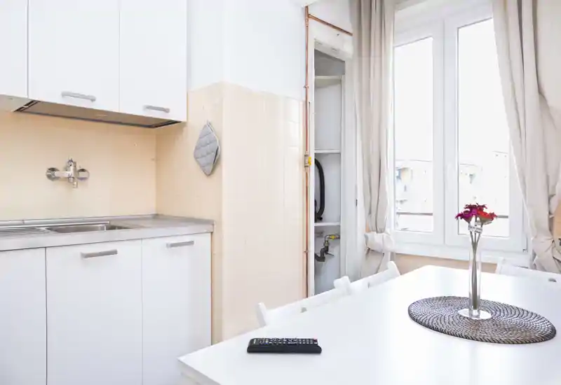 Bright, compact kitchen with white cabinets, stainless sink and a dining table by a large window providing generous natural light.