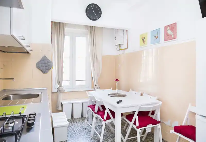 Bright, compact kitchen with a built-in sink and gas hob, a white dining table with red-cushioned folding chairs, and a window letting in natural light.