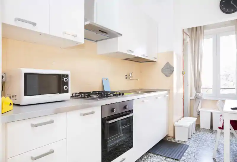 Bright, clean galley kitchen with built-in oven, gas hob and microwave; natural light from a large window and white cabinetry create a fresh, modern feel.
