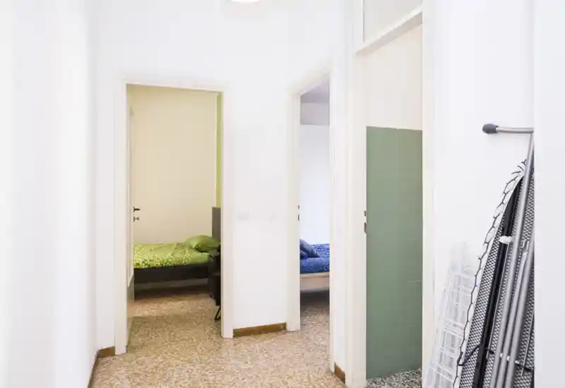 Hallway view showing two bedrooms through open doorways; tidy single beds with colorful bedding are visible, in a bright, minimally decorated apartment.
