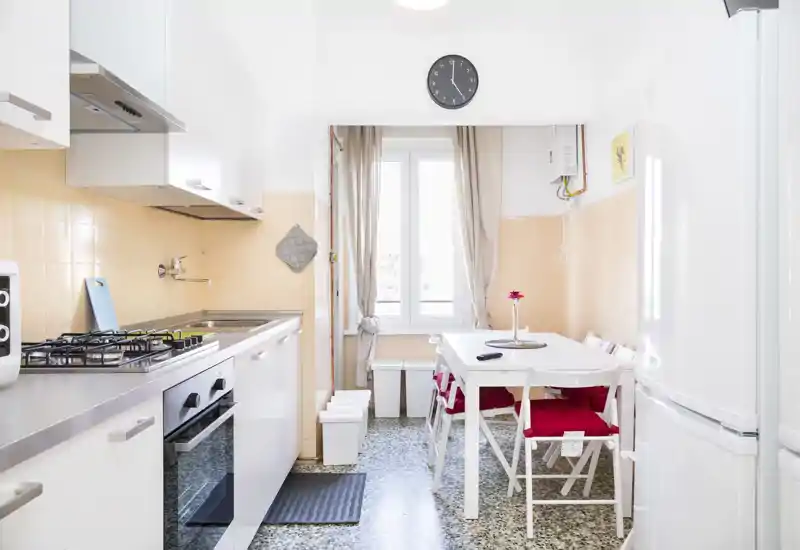 Bright, compact kitchen with a built-in oven and gas hob, a small dining table with red cushions and a window providing natural light — practical and well maintained for everyday use.