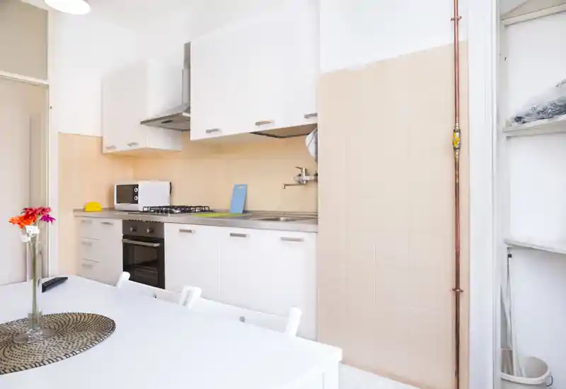 Bright, modern compact kitchen with white cabinetry, built-in oven and gas hob, stainless sink and a simple dining table in the foreground — neat and ready for use.
