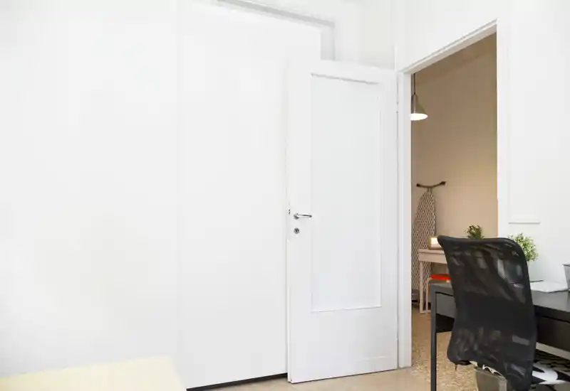 Bright, minimal room view showing a doorway, a desk with a mesh chair and a glimpse into an adjacent warmly lit space — suitable for a compact living or work area.