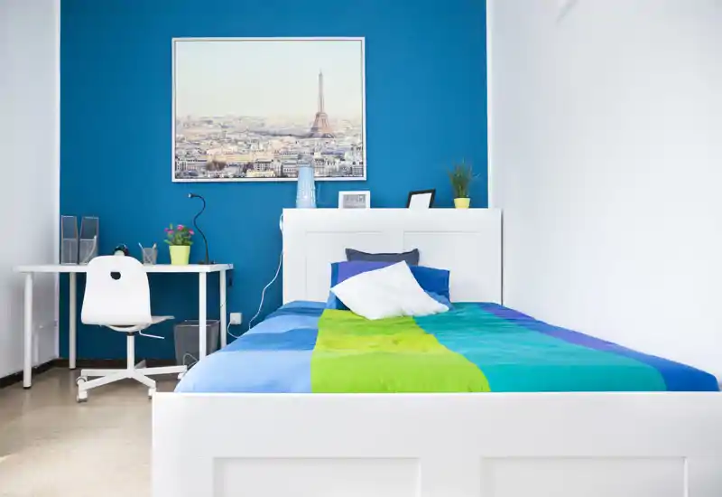 Camera da letto singola, luminosa e moderna con letto bianco, biancheria colorata e una piccola postazione di lavoro davanti a una parete d'accento blu — ideale per un annuncio che valorizza un ambiente accogliente e ordinato.