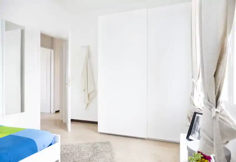 Camera da letto luminosa e minimalista con armadio bianco, tende chiare e una copertura letto colorata; ambiente pulito, arioso e ben illuminato, ideale per annunci.