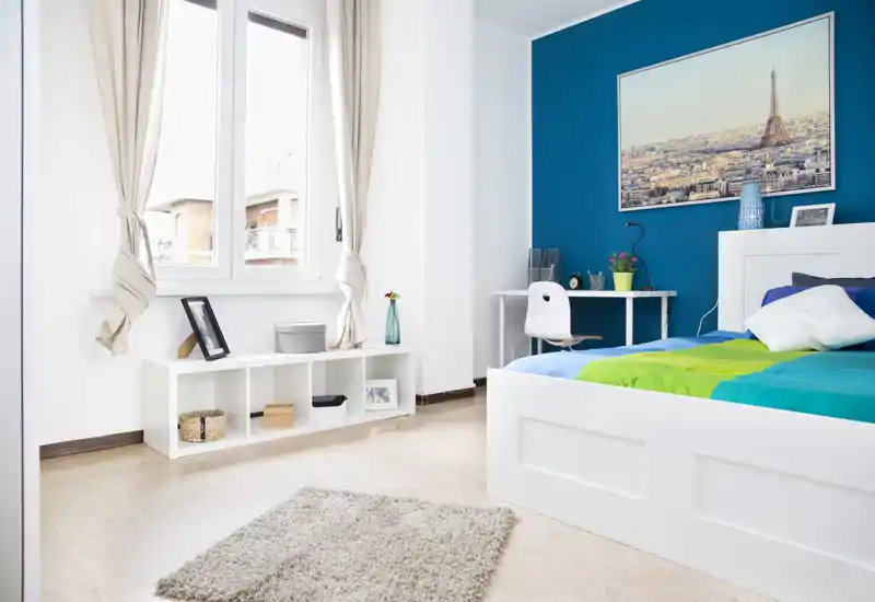 Camera da letto luminosa e moderna con letto piattaforma bianco, biancheria colorata e parete accentuata turchese; ampia finestra che illumina la stanza e una scrivania per lavorare.