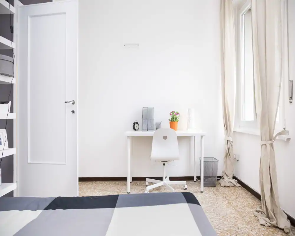 Bright, minimal bedroom with a visible double bed in the foreground, a simple desk by the window and soft natural light filling the room.