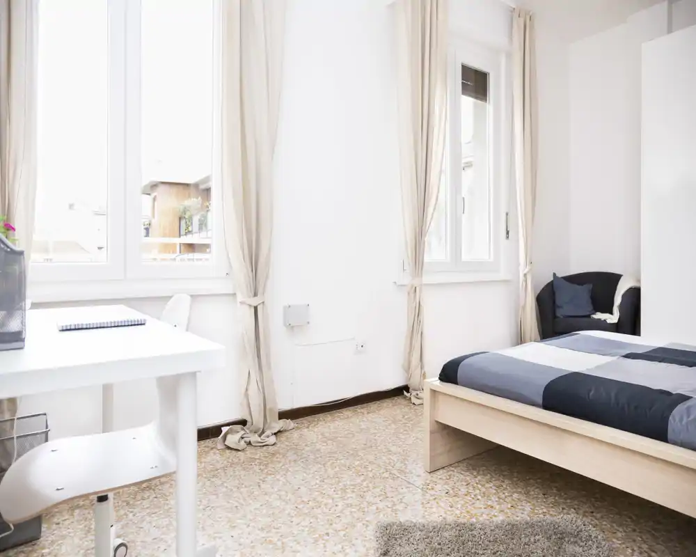 Bright, simply furnished bedroom with a double bed, a small desk by the window and a comfortable armchair — neutral linens and large windows create an airy, welcoming space.