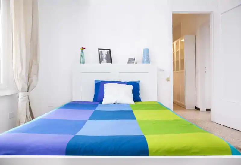 Chambre lumineuse et minimaliste avec une couette bleue et verte, tête de lit blanche et lumière naturelle provenant d'une fenêtre. Décoration simple avec un vase et des photos encadrées sur l'étagère de la tête de lit.