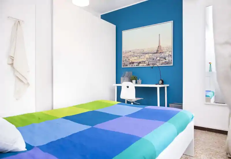 Chambre moderne et lumineuse avec un grand lit coloré, un petit bureau contre un mur accent bleu et une photo encadrée de la ville. Espace propre et bien éclairé, idéal pour voyageurs seuls ou location cosy.
