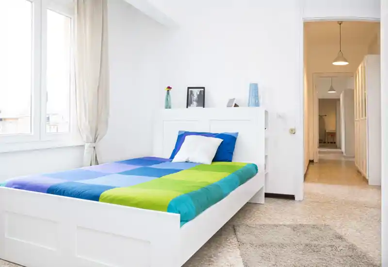 Chambre lumineuse et moderne avec un lit blanc habillé d'une couette bleu-vert, grande fenêtre apportant de la lumière naturelle et décoration minimale soignée.