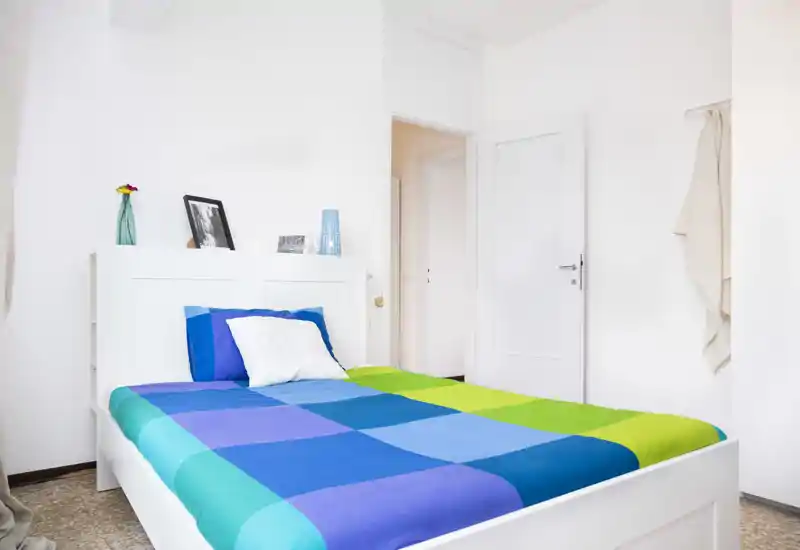 Chambre lumineuse et minimaliste avec un lit double coloré, une tête de lit blanche et des lignes modernes. La pièce est bien éclairée et décorée simplement avec quelques accessoires.