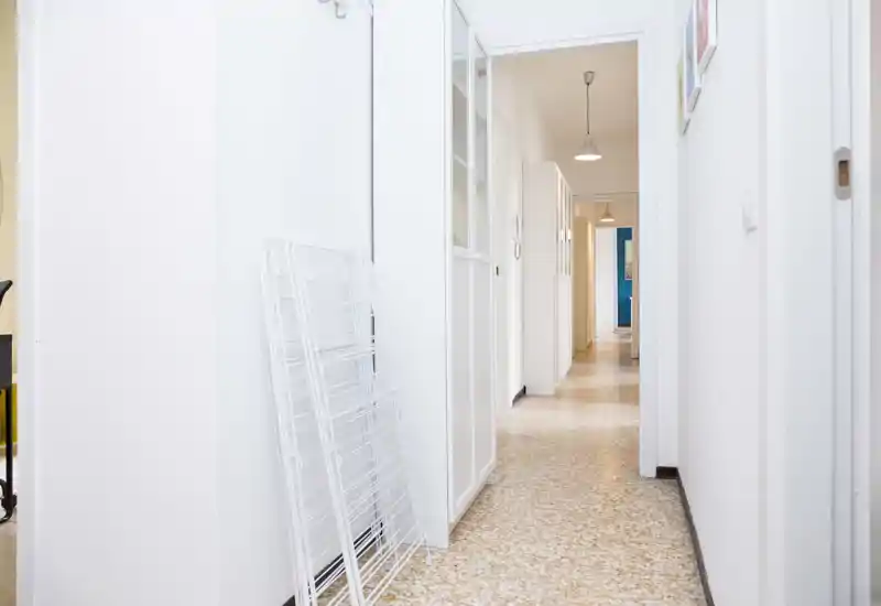Bright, clean hallway/entrance with terrazzo floor, white walls and pendant lights leading down a long corridor — practical entry area with storage and hooks.